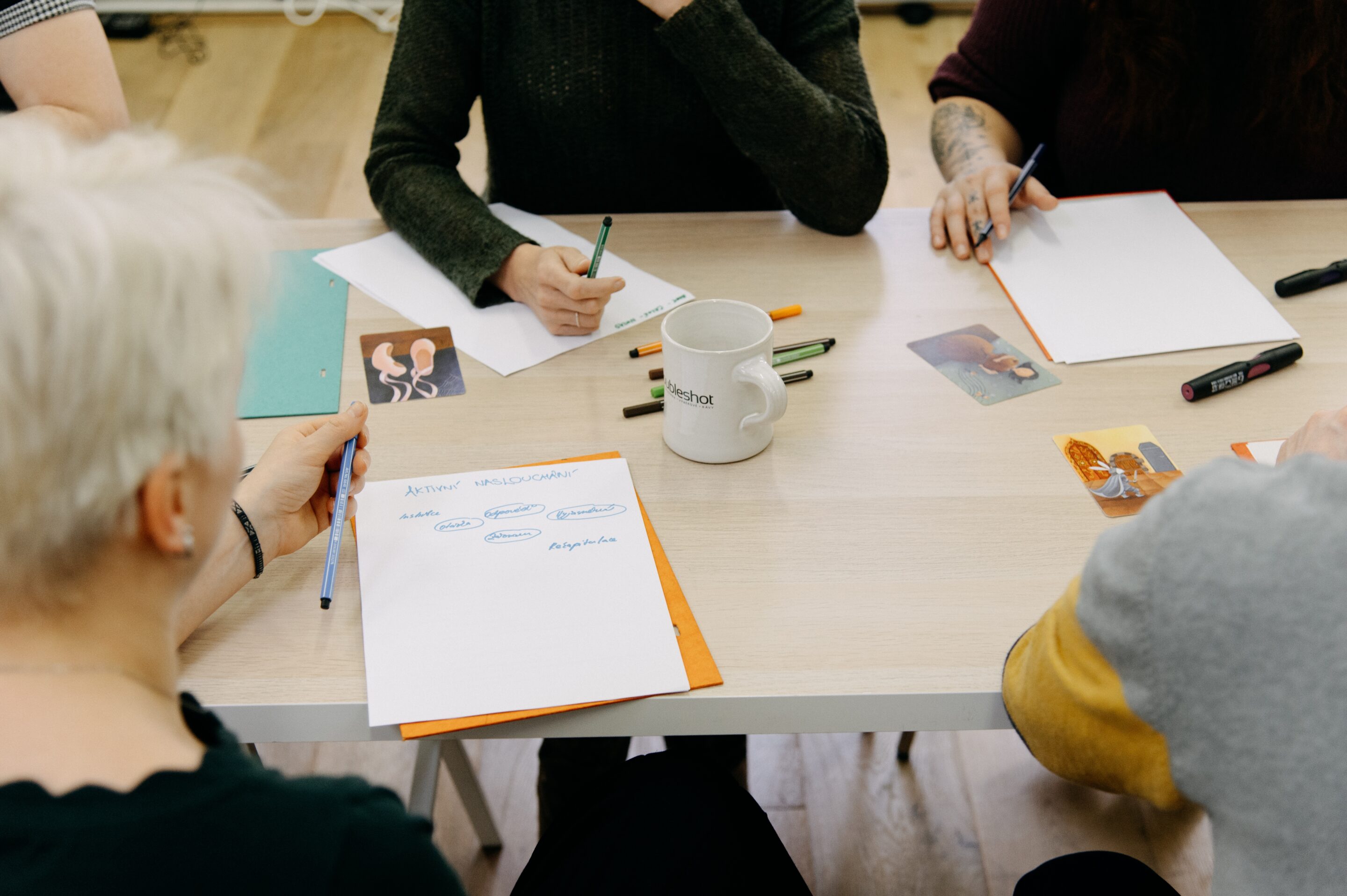 Pohled shora na stůl, kde účastníci kurzu Facilitační minimum pracují s papírem s nadpisem Aktivní naslouchání a rozebírají jednotlivé techniky vnímání skupiny.