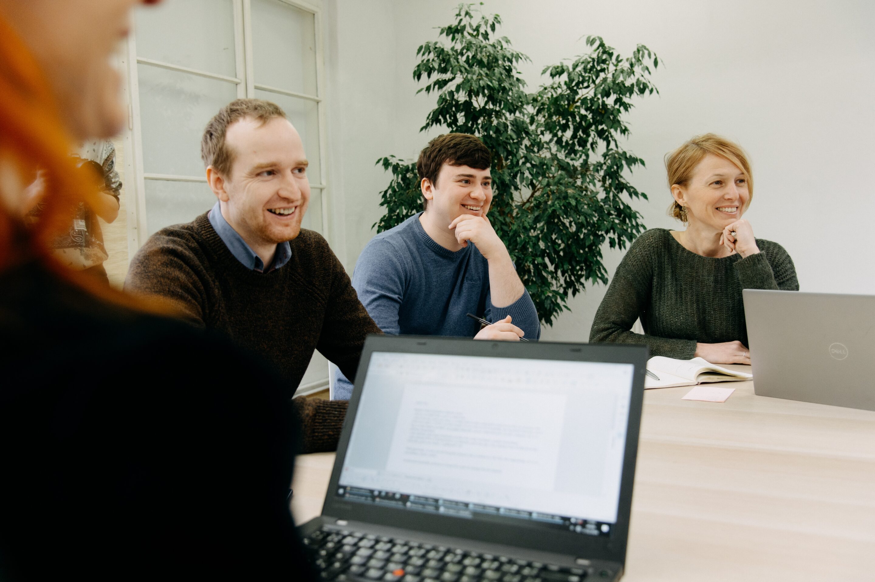 Tři usmívající se účastníci workshopu Edutica během diskuze u jednacího stolu s notebooky.